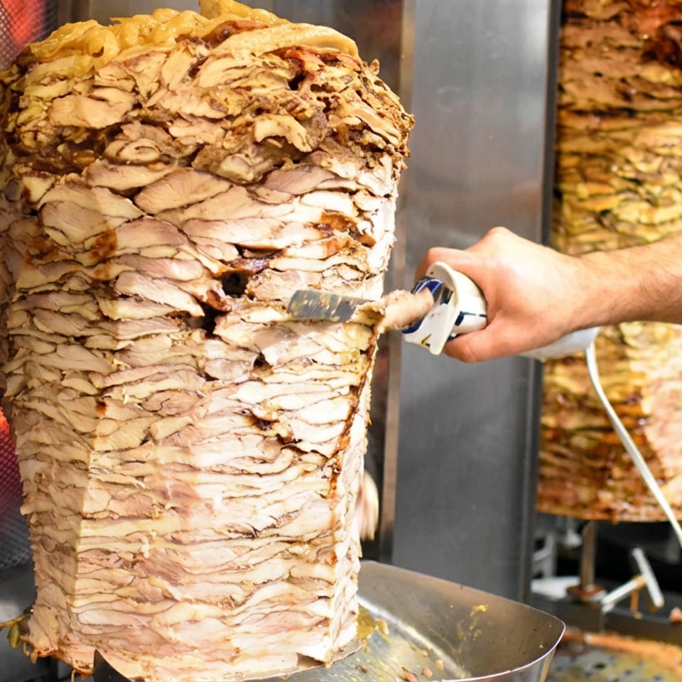 Worker slices a large vertical stack of roasted chicken shawarma beside additional rotating meat spits.